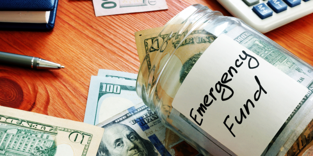 Opened jar that says "emergency fund" is on its side on desk with U.S. cash inside and around it.