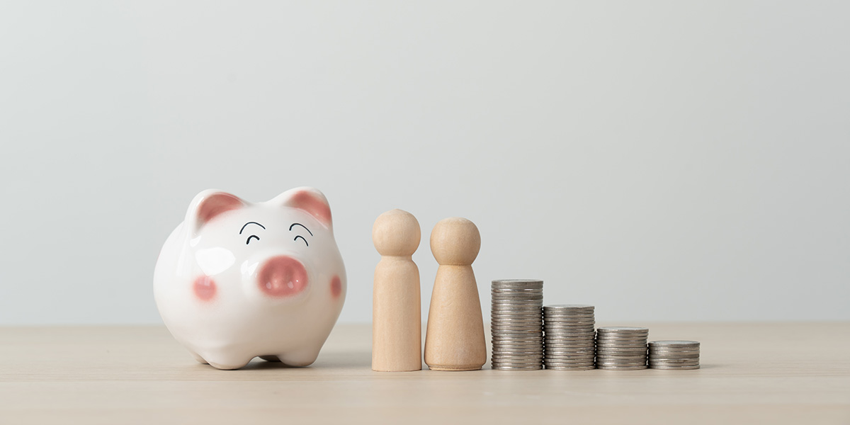 a pink and white piggy bank sits at the head of a line to two wooden figurines and four piles of coins. they sit on a light wood grain surface with a white background